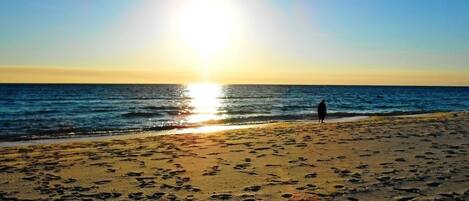 Una playa con huellas que conducen al agua, una persona de pie cerca de la orilla y el sol poniéndose en el horizonte.