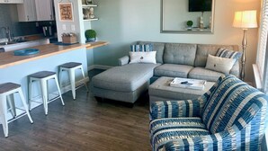 Un salón moderno con un sofá gris, un reposapiés a rayas y una zona de cocina con armarios blancos y encimera de madera.