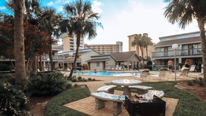 Zona de piscina con sillones de descanso y una parrilla, rodeada de palmeras y un edificio con balcones.