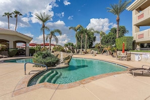 A pool area with clear water, lounge chairs, palm trees, and a building with a red roof.