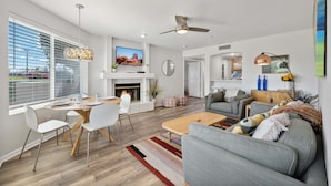 A modern living room with a fireplace, a dining area with a table and chairs, a grey sofa, and a ceiling fan.