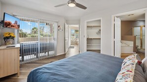 A bedroom with a bed, a ceiling fan, a TV, and a view through a window.