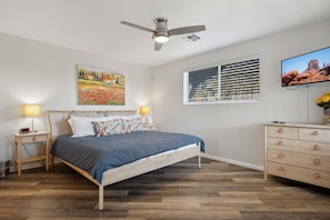 A bedroom with a bed, bedside lamps, a ceiling fan, a window with blinds, a dresser, and a TV on the wall.