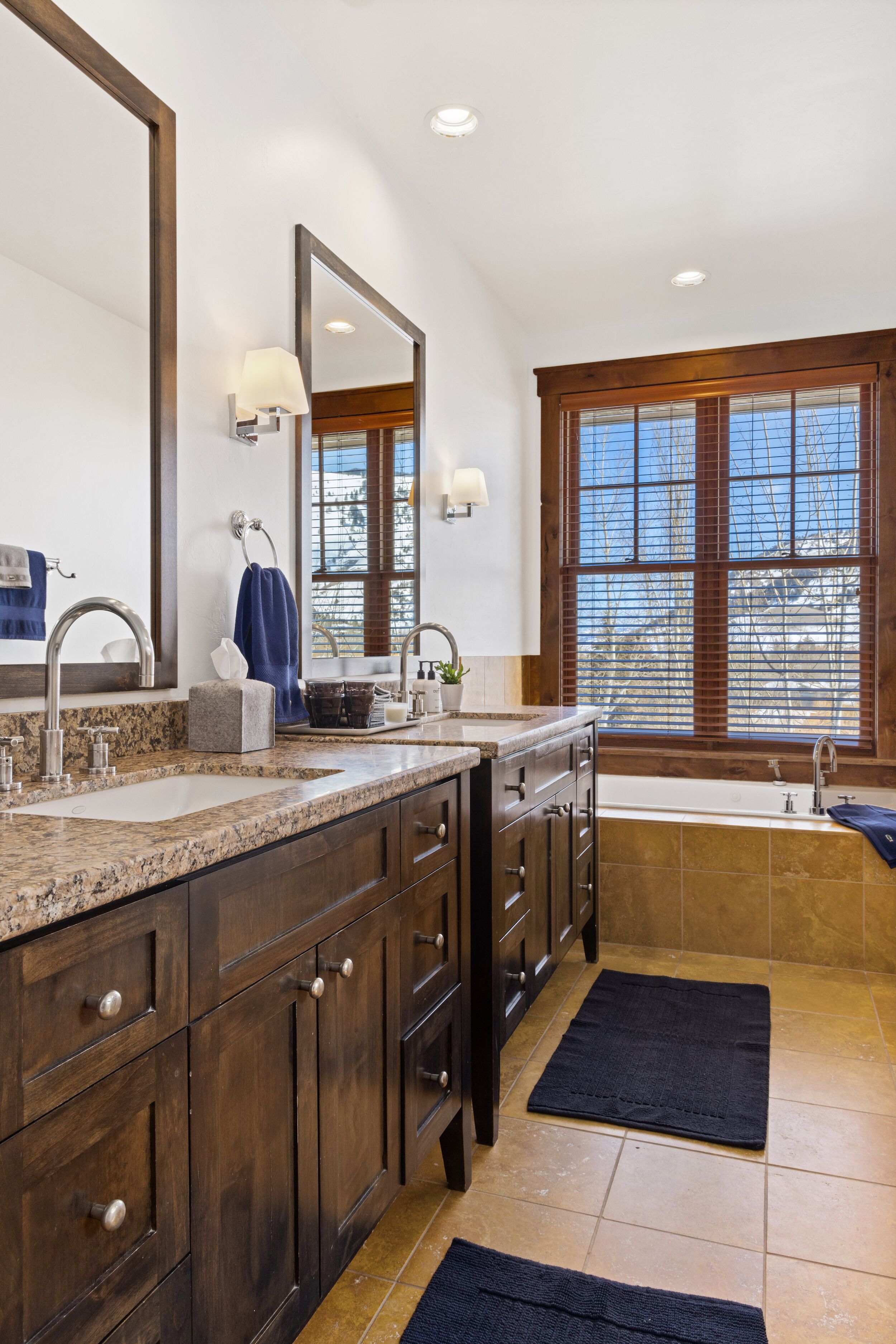 A modern bathroom with a large vanity, double sinks, a bathtub, and a window with blinds.