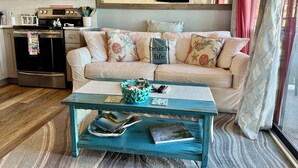 A cozy living room with a beige sofa, teal coffee table, and a kitchen area with stainless steel appliances. - Stunning Oceanview Luxury Remodel, Bikes Included! (Hilton Head Island)