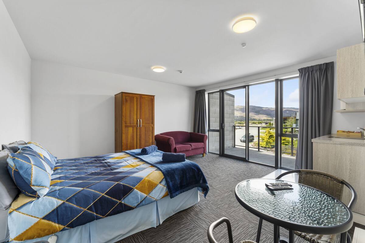 A hotel room with a bed, a red armchair, a glass table, and a view of the outdoors.