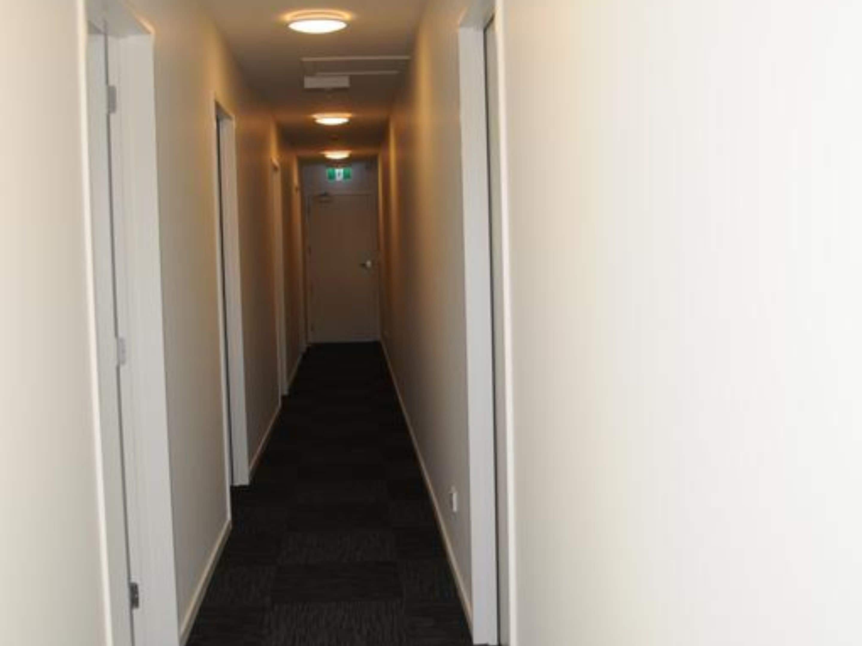 Un couloir étroit, recouvert de moquette, avec des murs et des portes blanches de chaque côté, qui mène à une porte au bout.
