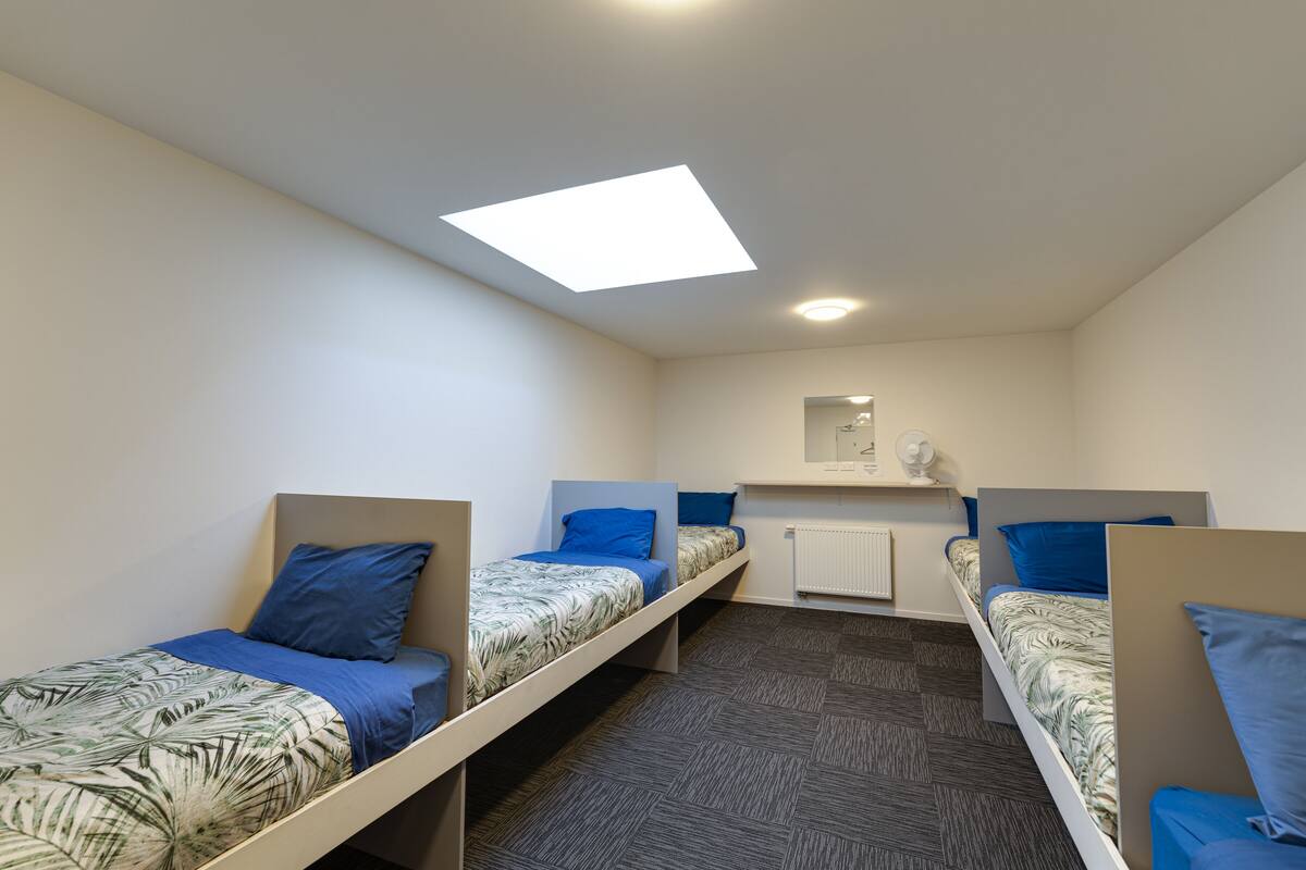 A dormitory room with three single beds, each with a blue pillow and a patterned bedspread. There is a skylight above the beds and a white radiator on the wall.