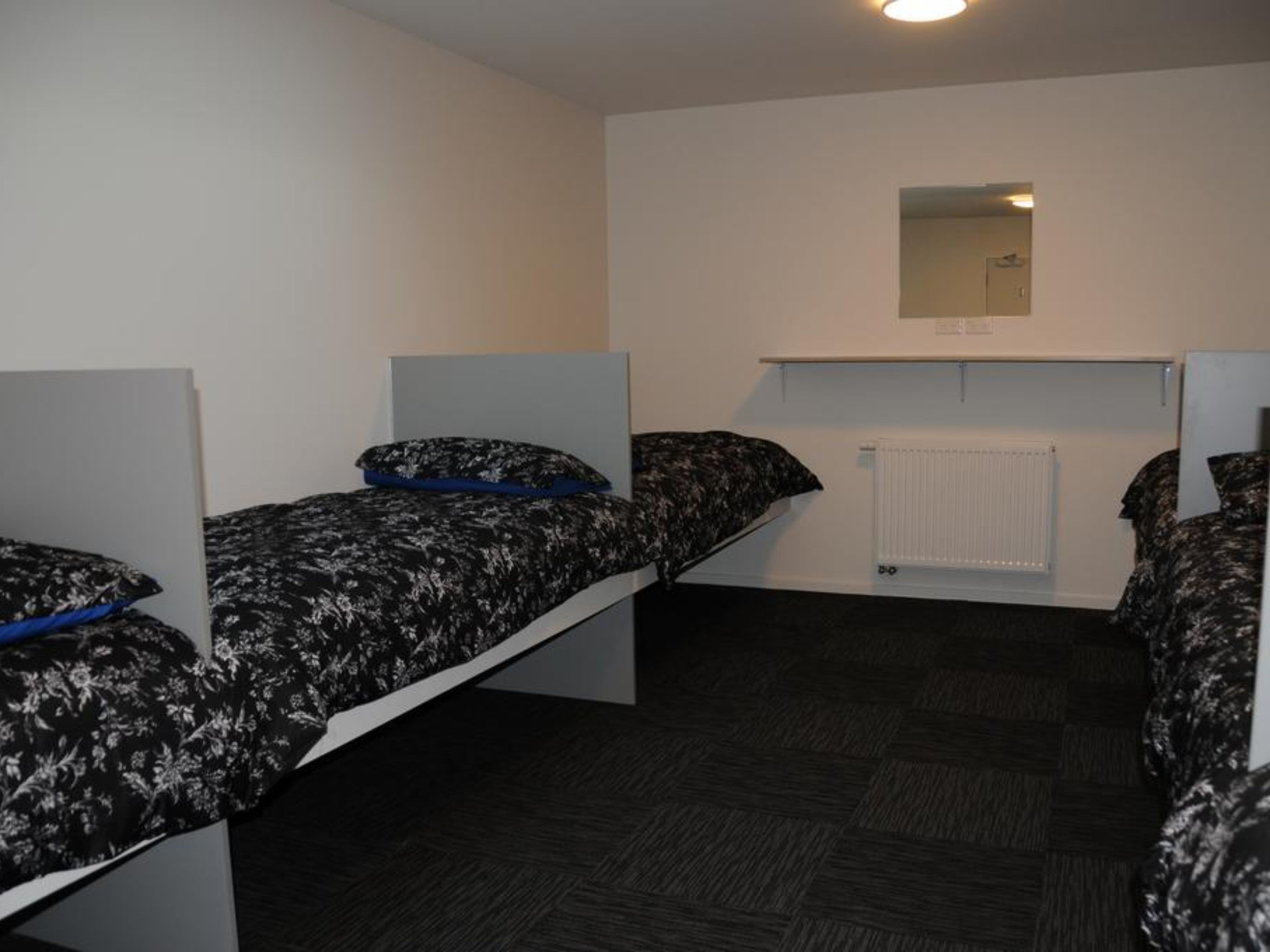 A dormitory room with two single beds, each with a patterned bedspread and pillows. There is a white radiator below a mirror on the wall, and the floor is covered with dark carpet.