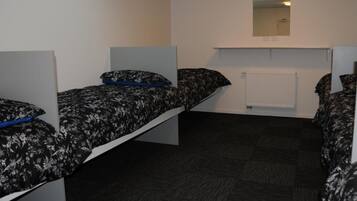 A dormitory room with two single beds, each with a patterned bedspread and pillows. There is a white radiator below a mirror on the wall, and the floor is covered with dark carpet.