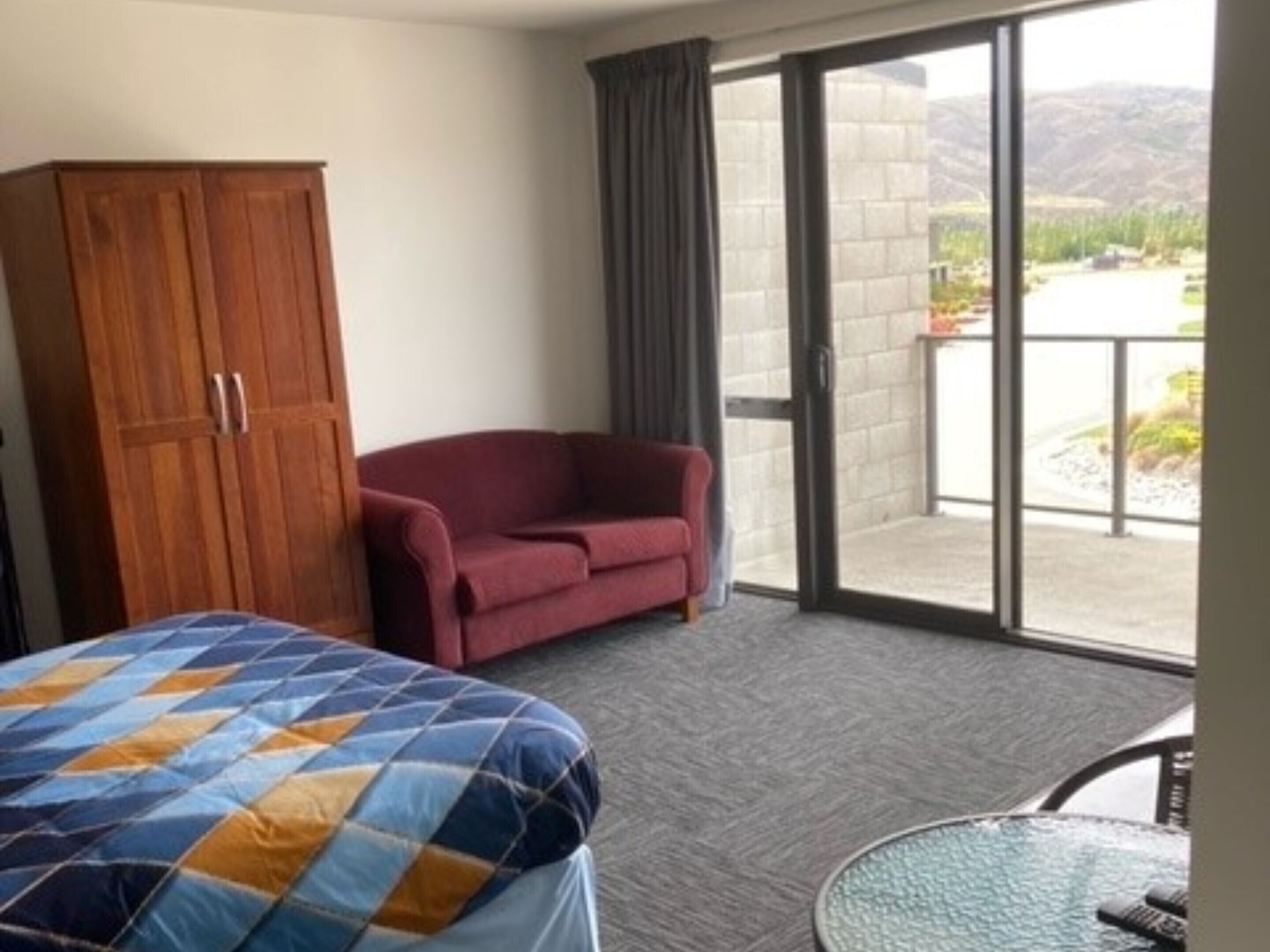 Een hotelkamer met een bed, een rode fauteuil, een houten garderobe en een glazen schuifdeur naar een buitenruimte.