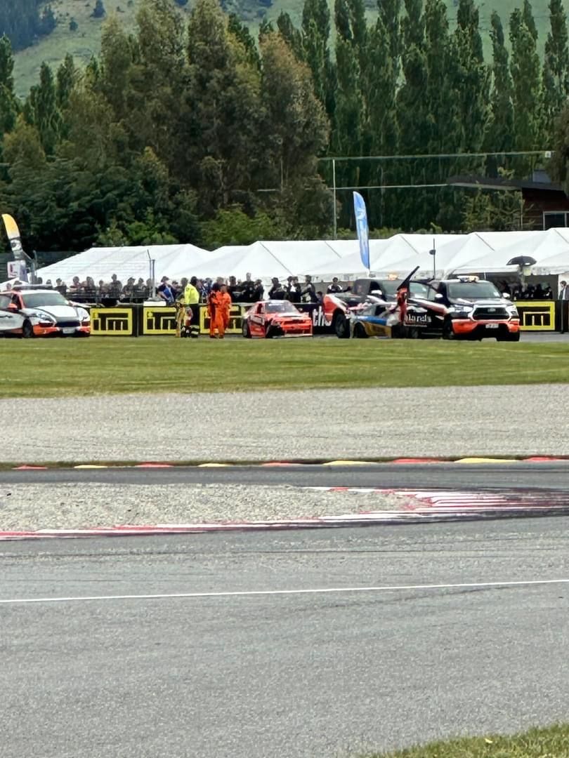 A race track with multiple cars lined up, spectators, and tents in the background.