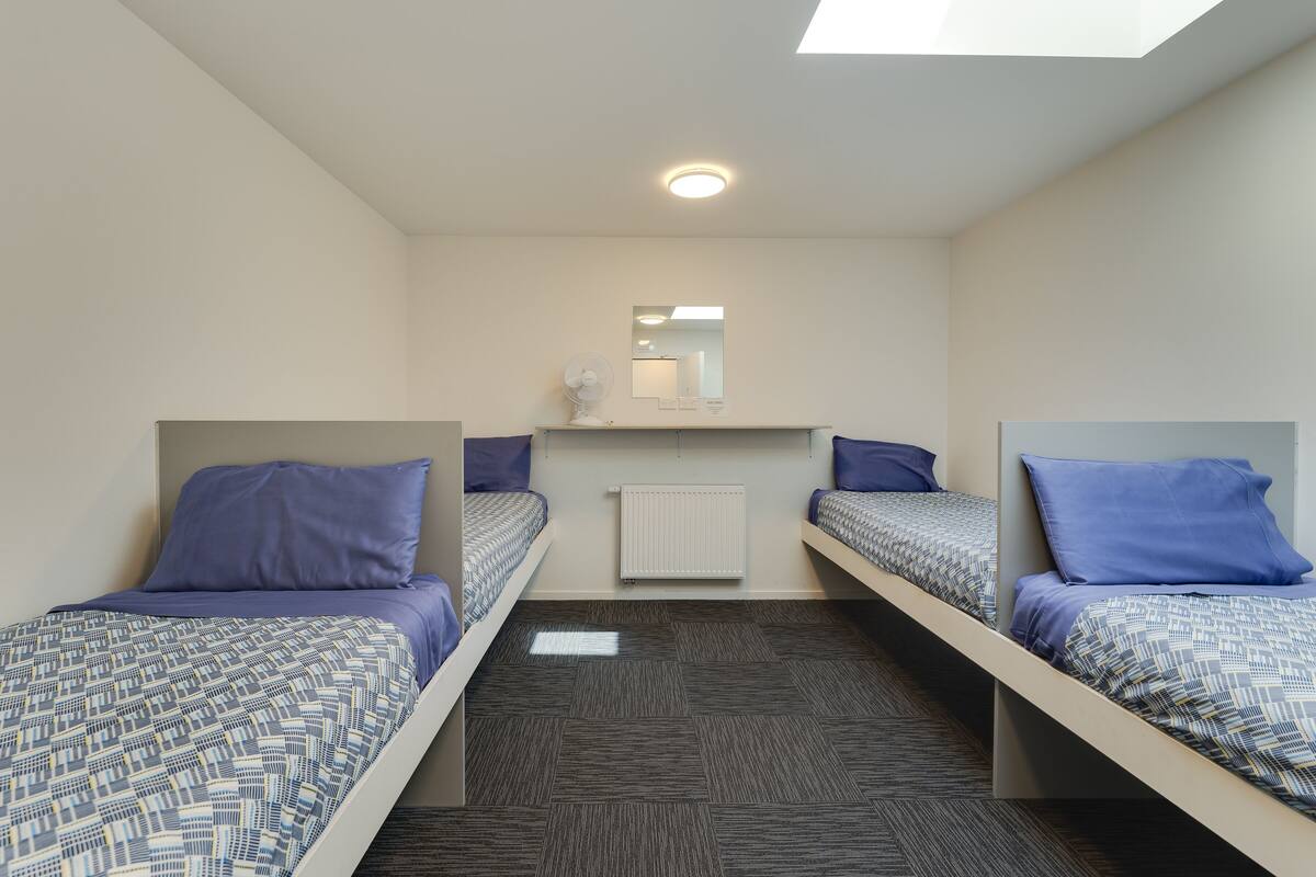 A room with three single beds, each with a patterned bedspread and matching pillows. There is a ceiling fan, a radiator, and a mirror above the beds.
