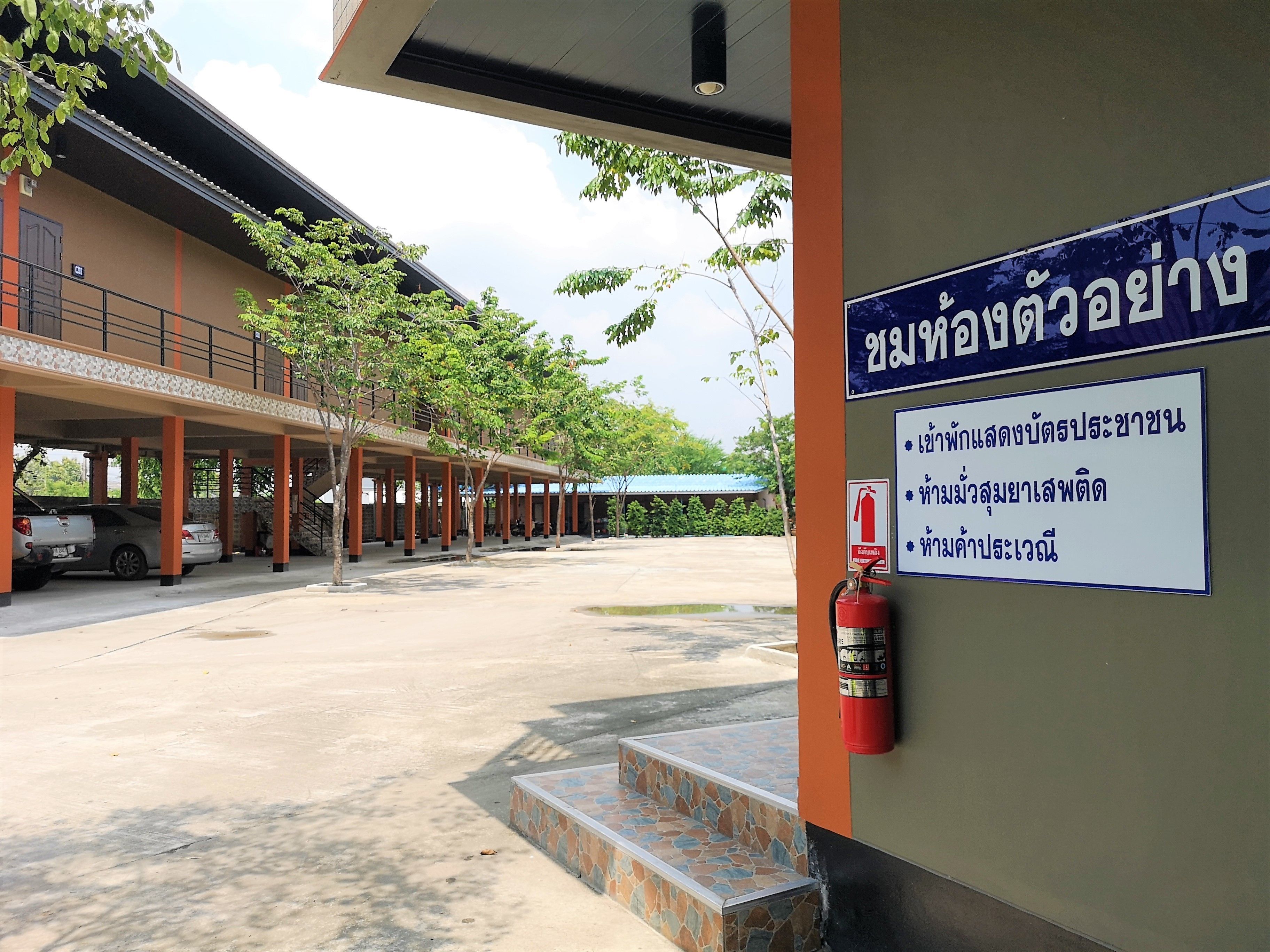 A covered parking area with a fire extinguisher mounted on the wall, a staircase leading to an upper level, and a sign with Thai text.