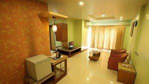 A hotel room with a kitchenette, a seating area with a sofa and armchairs, a small table with a lamp, and a window with curtains. - Hotel Kohinoor Square (Kolhapur)