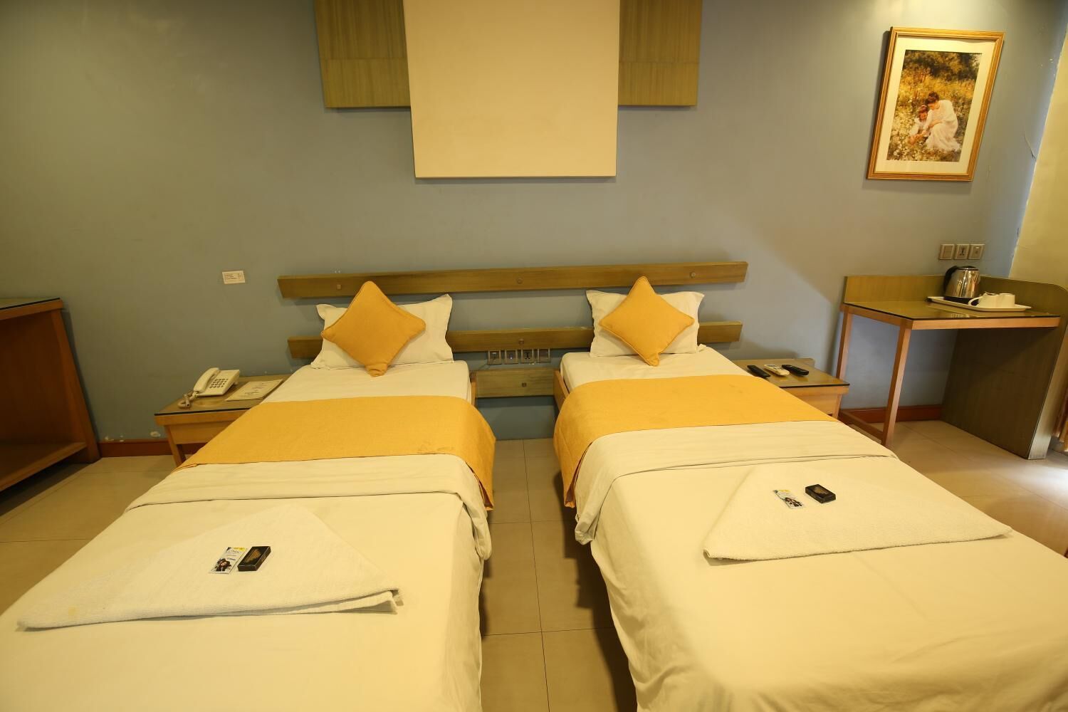 A hotel room with two beds, a wooden headboard, a bedside table with a phone, a small table with a kettle and cups, and a painting on the wall.