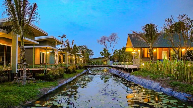 A canal with water lilies, surrounded by buildings with warm lighting, and palm trees. - Green 2 (Chanthaburi)