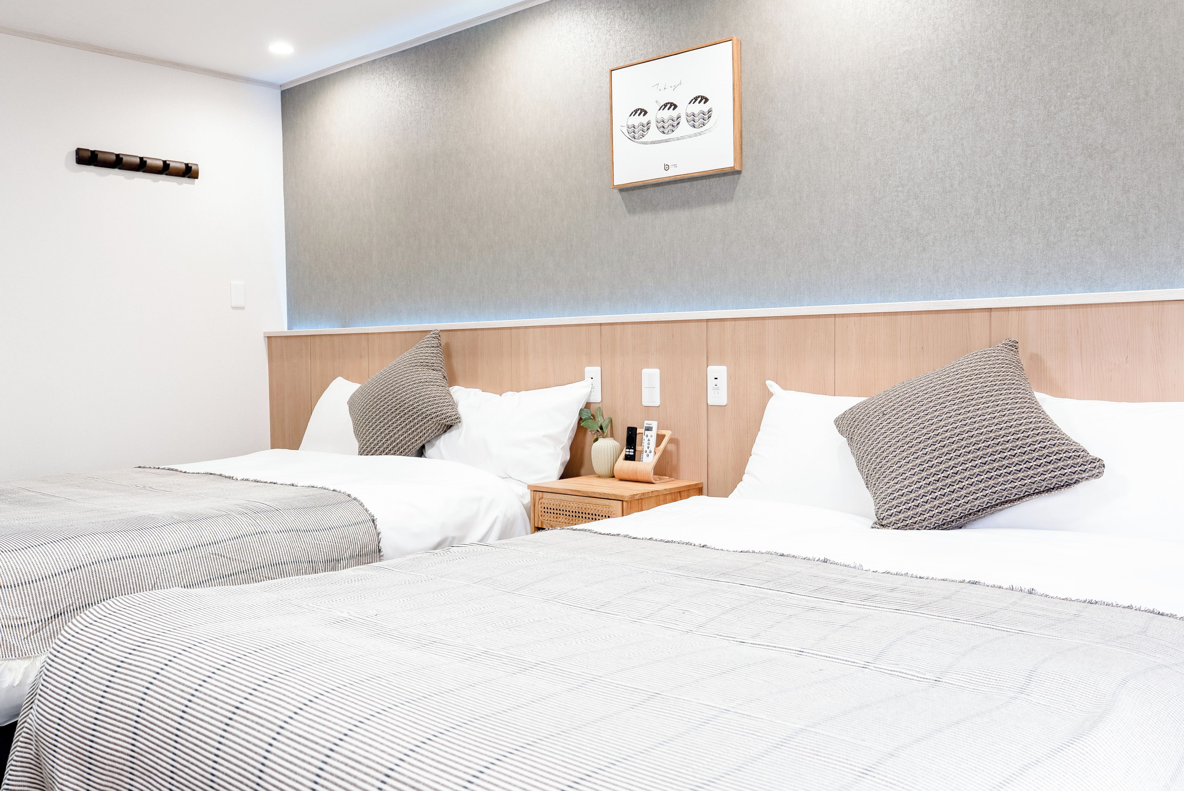 A hotel room with two beds, a bedside table with a phone and a plant, and a framed artwork on the wall.