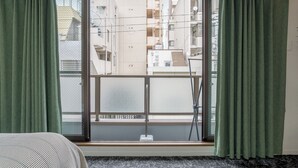 Una habitación con puerta corrediza de vidrio que da a un balcón, una cama con colcha a rayas y una ventana con cortinas verdes.