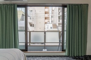 A room with a sliding glass door leading to a balcony, a bed with a striped bedspread, and a window with green curtains. - b&Dotonbori 3 (Osaka)