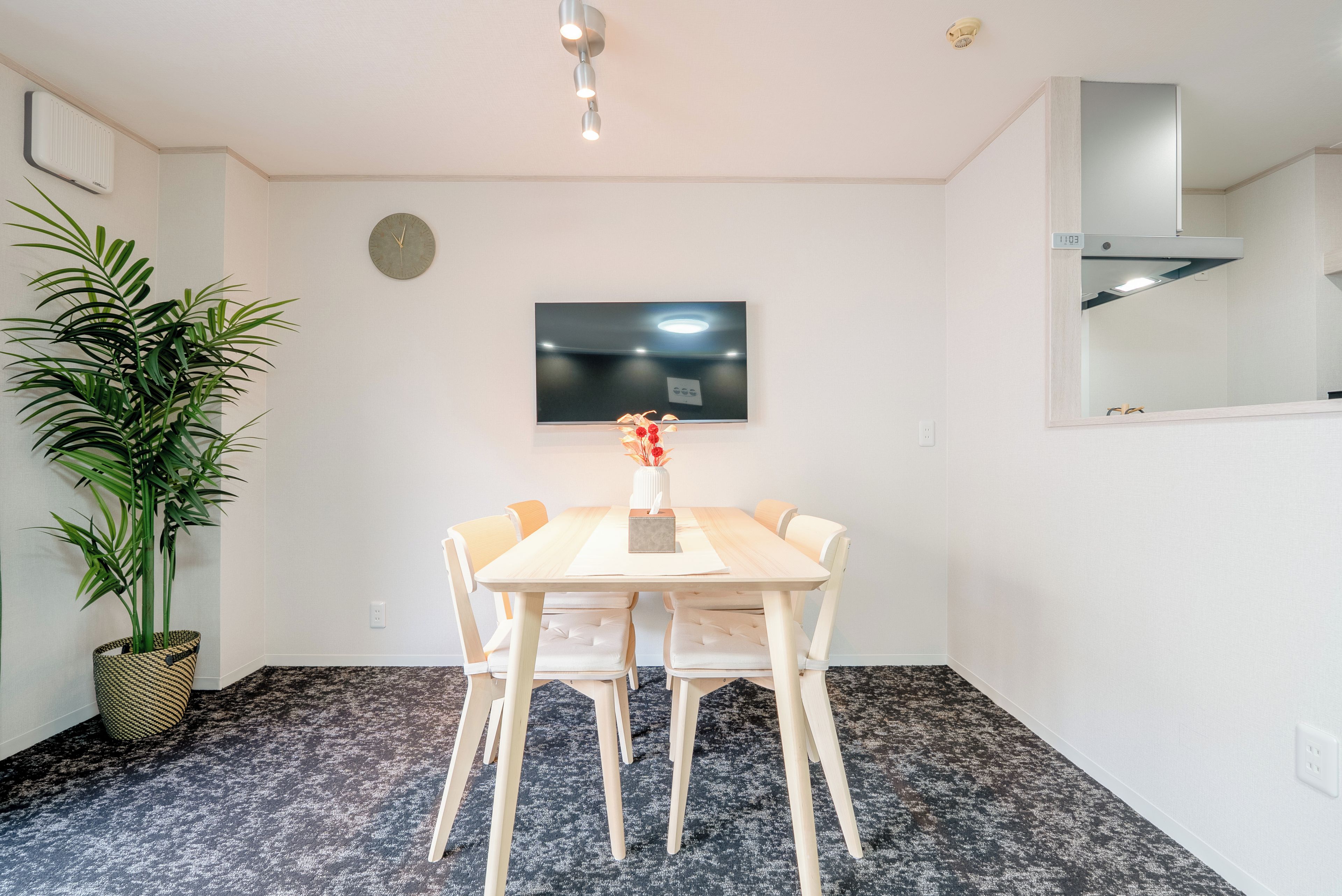 A dining area with a table and chairs, a potted plant, a wall clock, and a flat-screen TV mounted above the table.