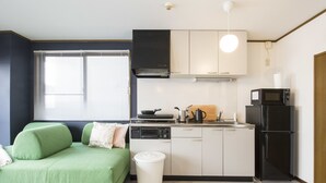 A compact kitchen with a stove, microwave, and refrigerator. There is a green sofa and a window with blinds.