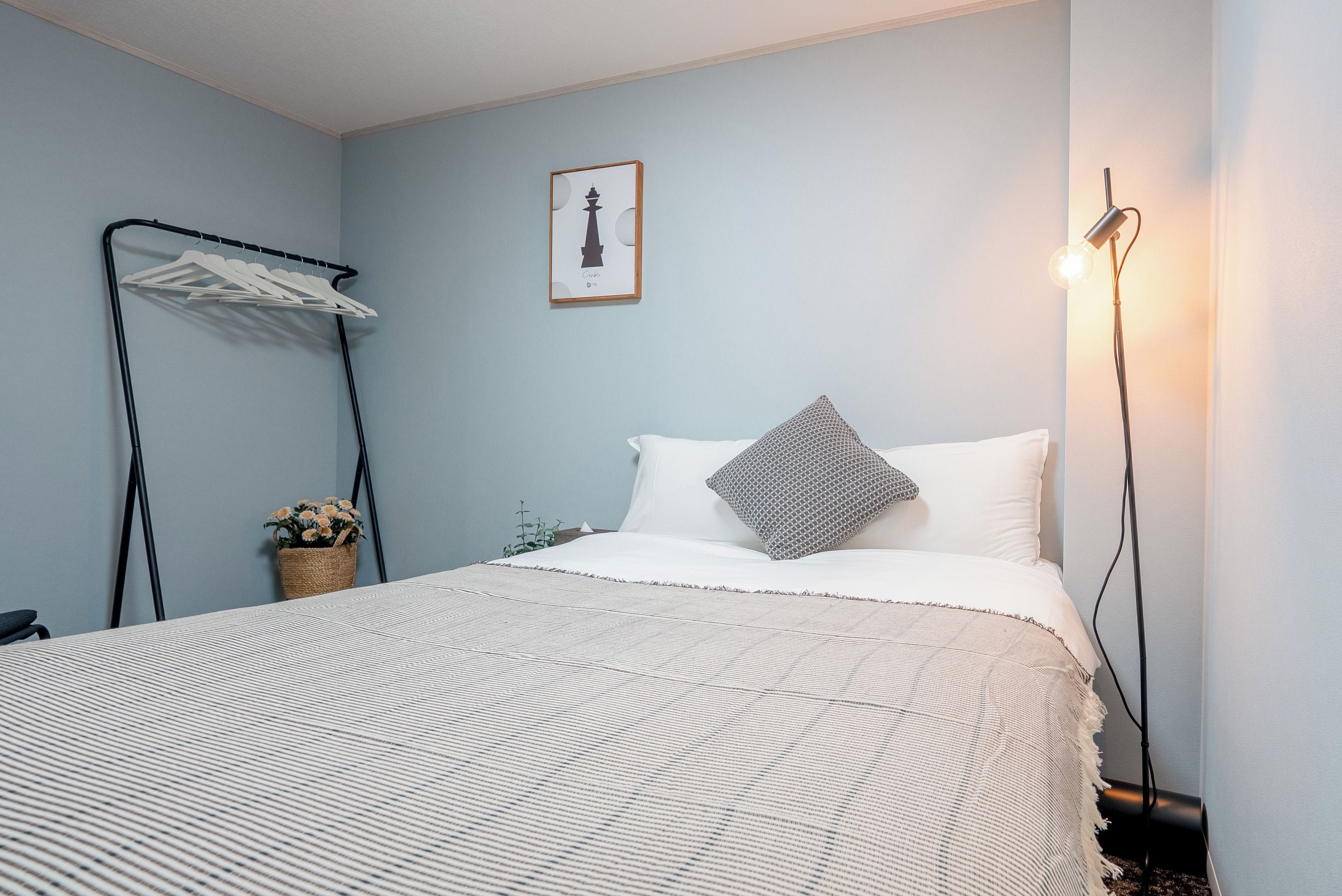 A neatly arranged bedroom with a bed, a hanging clothes rack, a framed picture on the wall, and a bedside lamp.