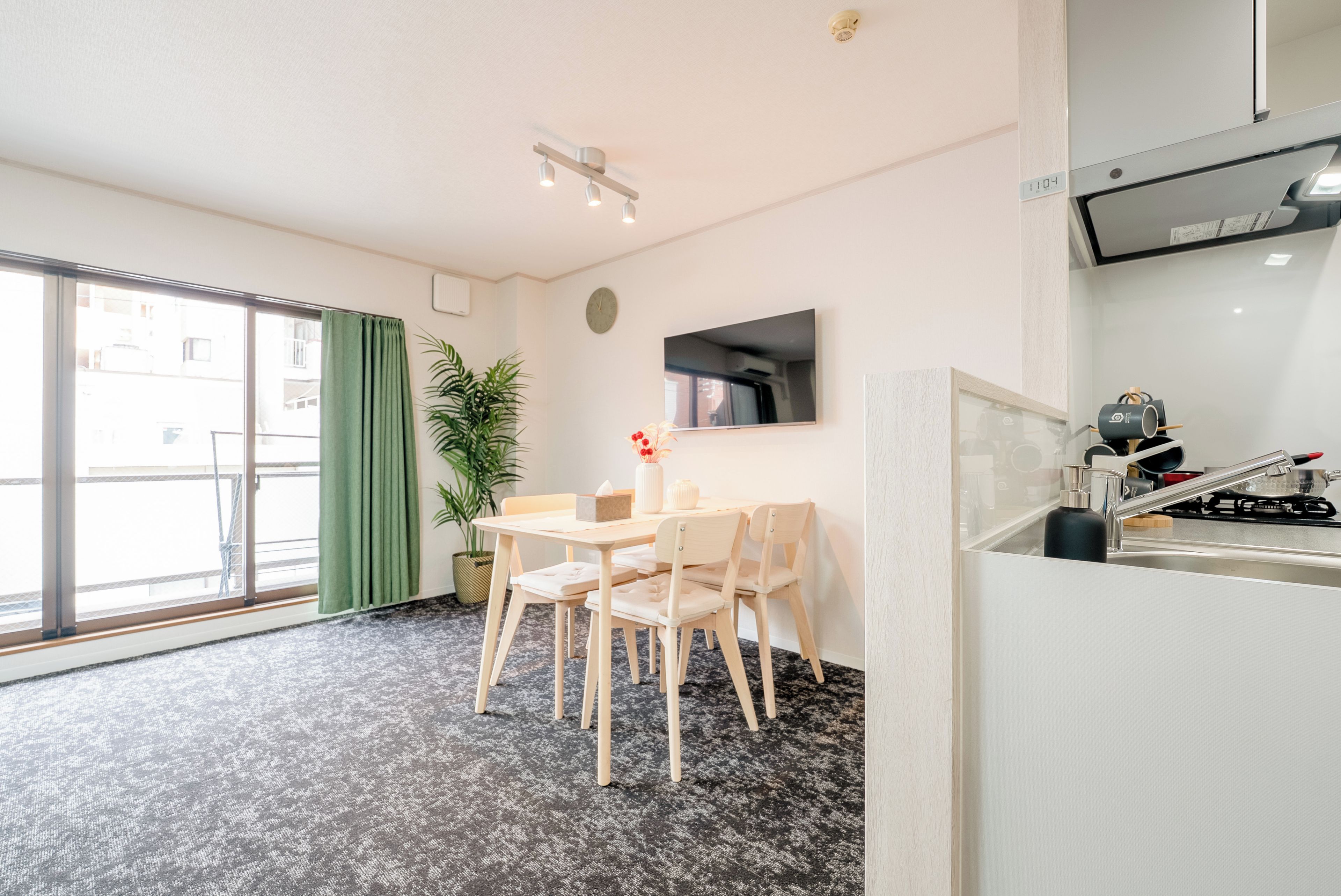 A hotel room with a dining table, chairs, a potted plant, a flat-screen TV, and a kitchenette.