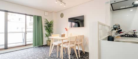 A hotel room with a dining table, chairs, a potted plant, a flat-screen TV, and a kitchenette.