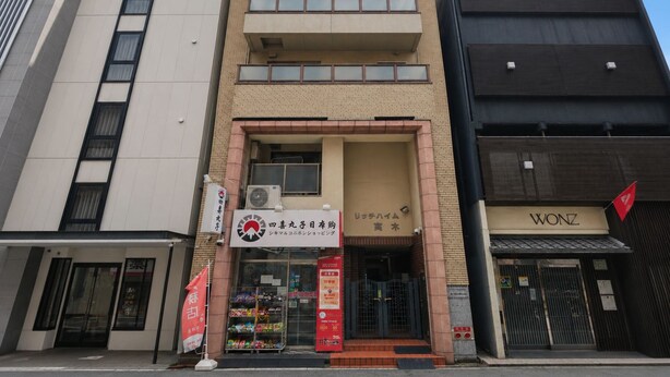 Un edificio de varios pisos con entrada a nivel de calle, una tienda visible con letrero rojo, y un edificio negro con una bandera roja a la derecha.