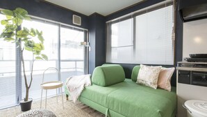 A compact living space with a green sofa, a small round ottoman, a potted plant, and a kitchenette area.