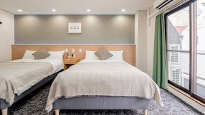 Habitación de hotel con dos camas, una mesita de noche con lámpara, aire acondicionado de pared y una ventana con vistas a edificios.