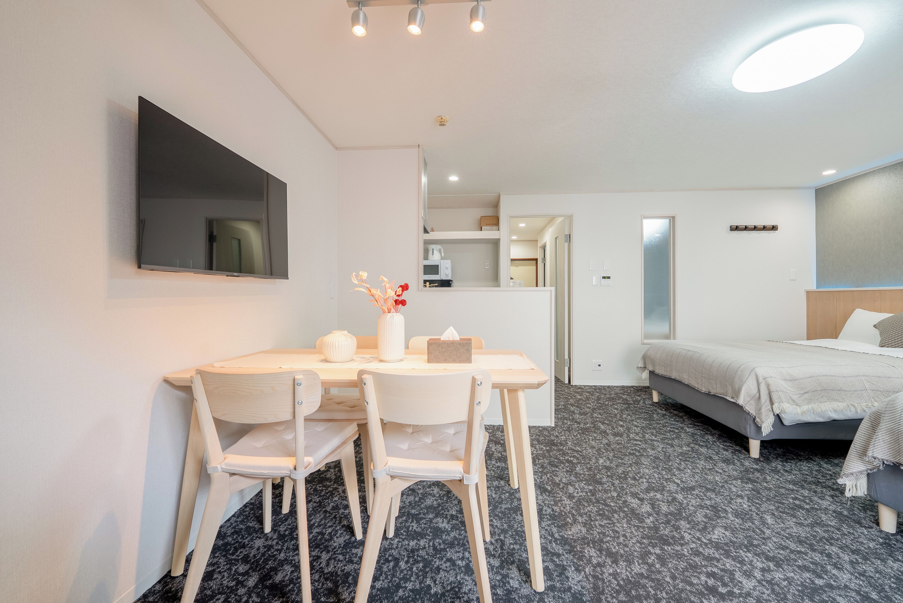 A hotel room with a bed, a desk with chairs, a flat-screen TV, a mirror, and a kitchenette area.