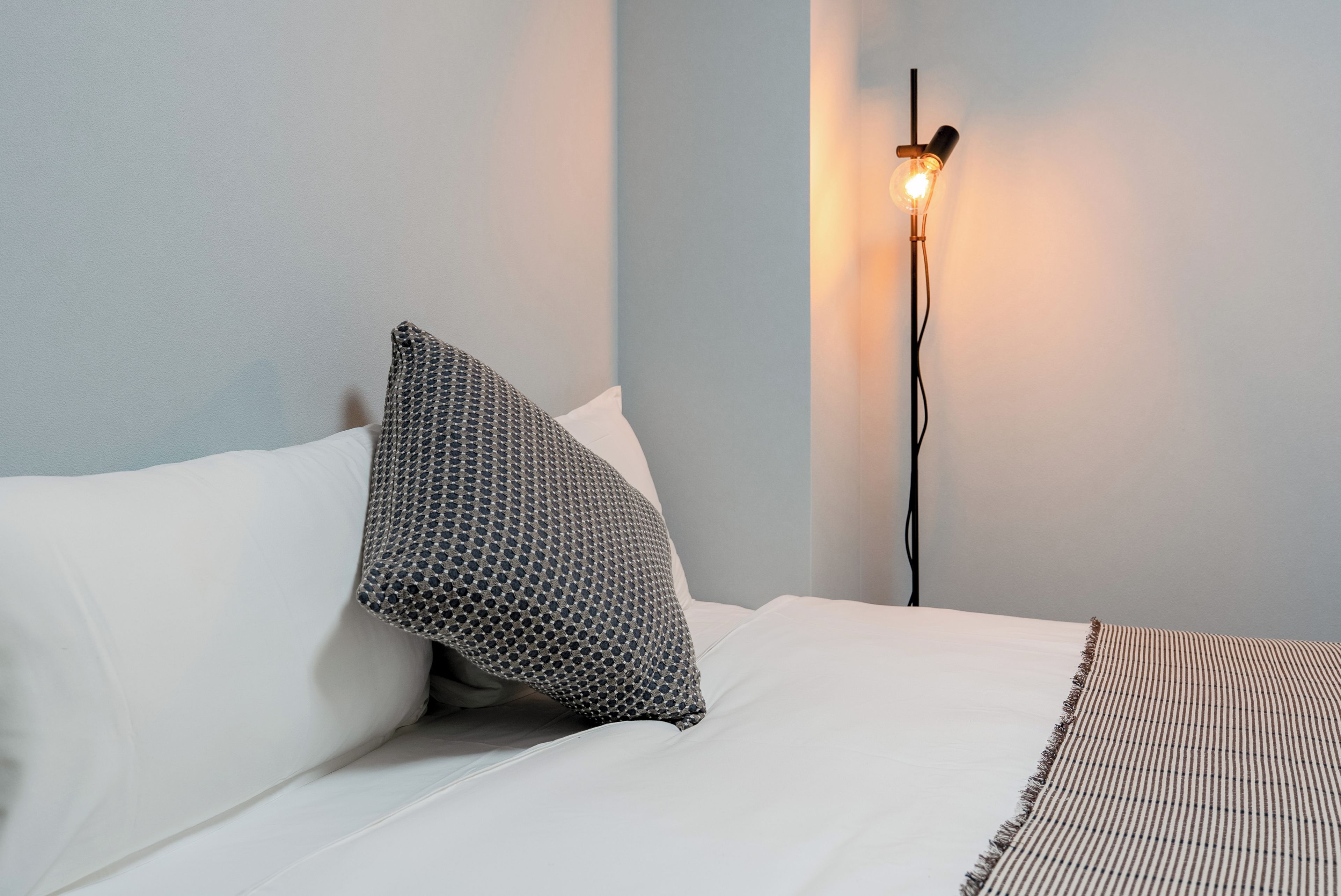 Una cama con ropa blanca y una almohada estampada, una lámpara de noche y una manta a rayas.