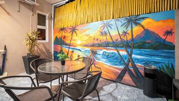 An outdoor seating area with a round table and chairs, a potted plant, and a mural of a sunset over the ocean with palm trees.