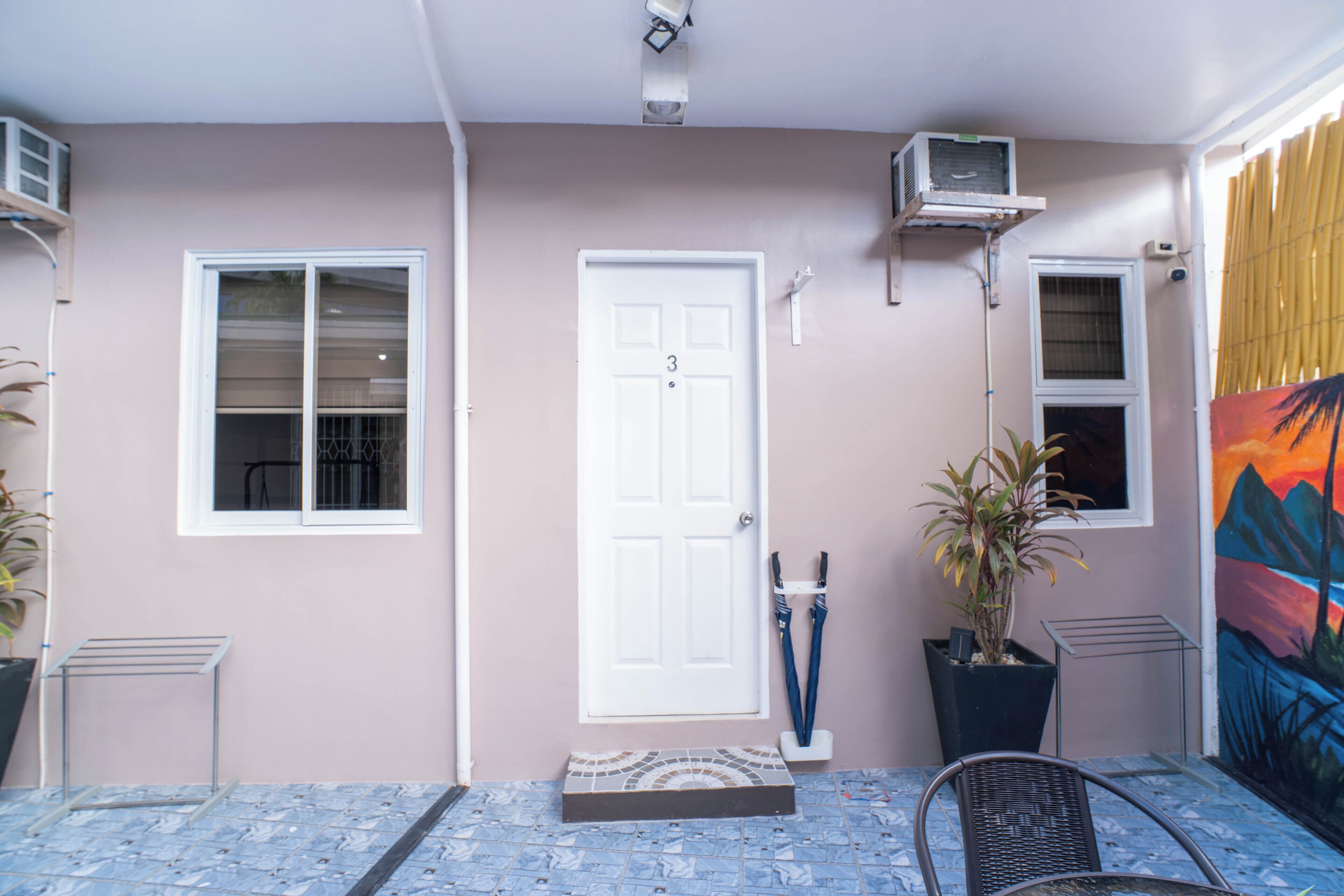 A covered outdoor area with a white door, two windows, a potted plant, a metal chair, and a tiled floor.