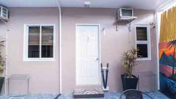 A covered outdoor area with a white door, two windows, a potted plant, a metal chair, and a tiled floor.