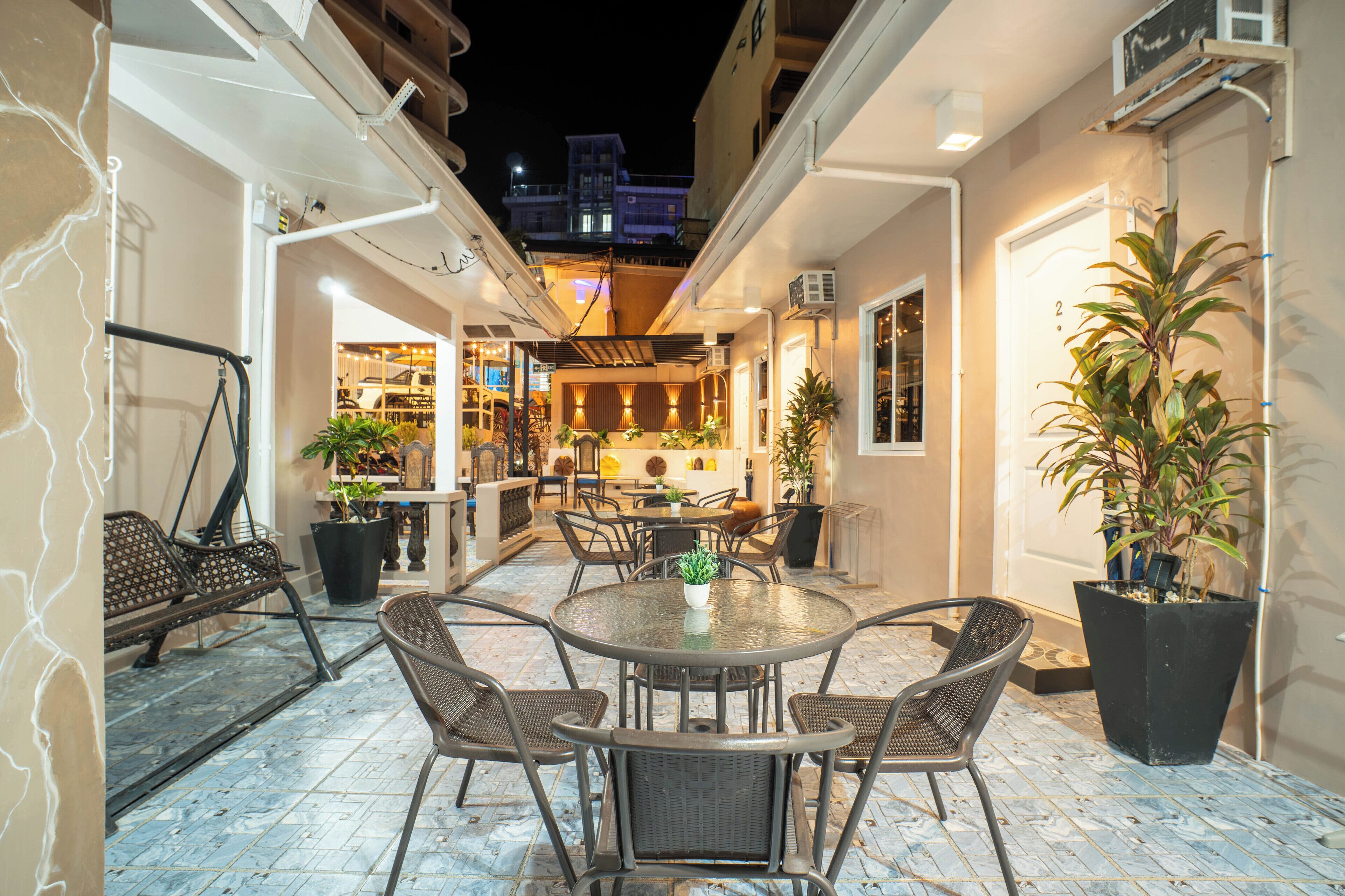 An outdoor patio area with a round table and chairs, potted plants, and a view of a building in the background.