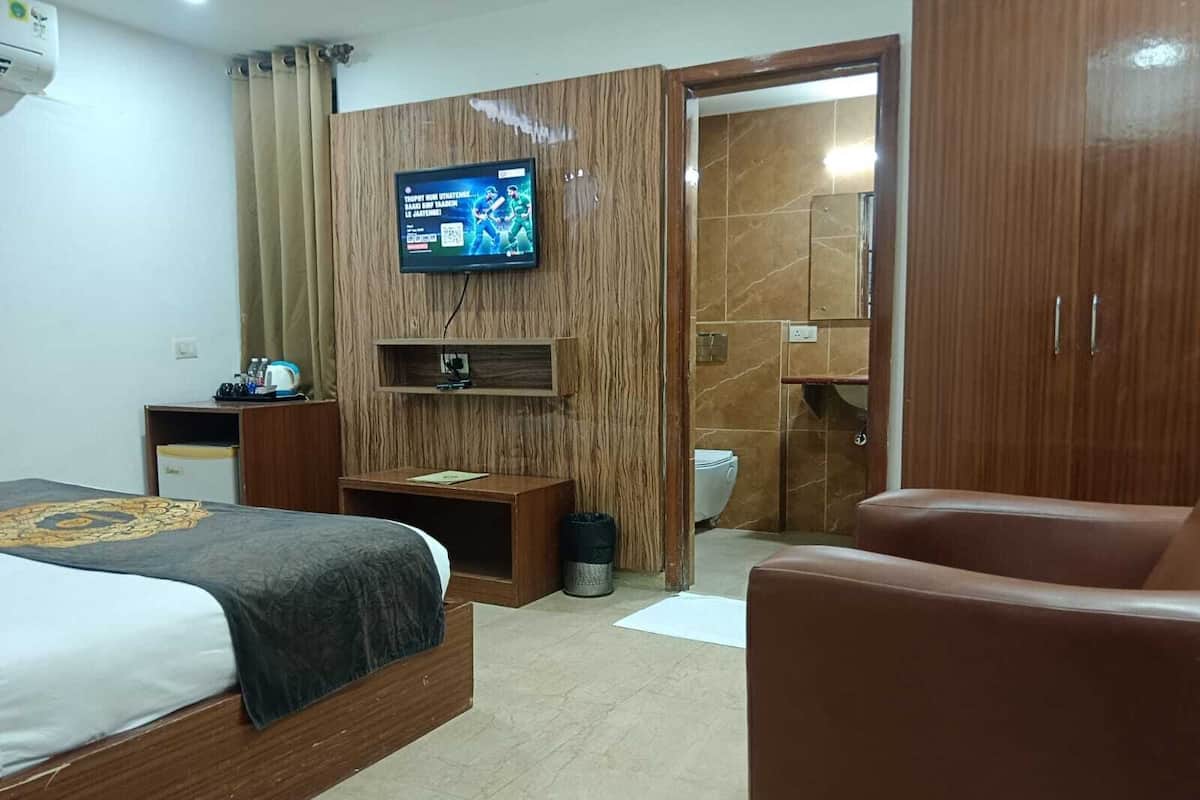 A hotel room with a bed, a TV mounted on the wall, a wooden wardrobe, a small table, and a bathroom visible through an open door.