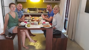 Um grupo de pessoas sentadas em uma mesa de madeira em uma cozinha, com uma geladeira, pia e diversos alimentos sobre o balcão.