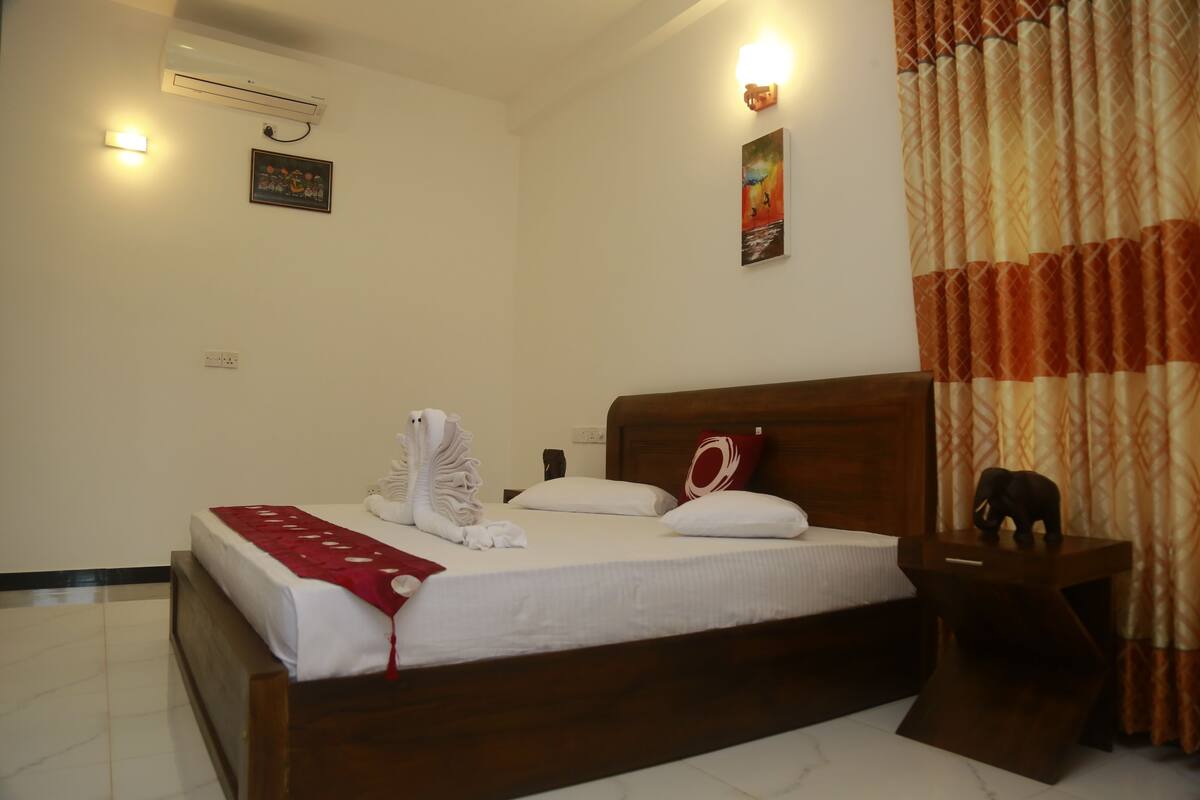Una habitación de hotel con una cama de madera, ropa de cama blanca, un plaid rojo, un aire acondicionado de pared, una luz de noche y una mesita de noche de madera con una estatua de elefante decorativa.