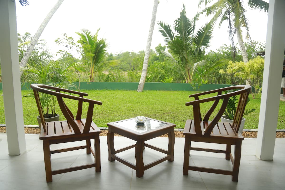 Un patio con dos sillas de madera y una mesa de vidrio, con vista a un jardín exuberante con palmeras y una cerca verde.