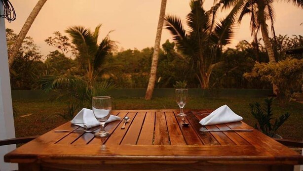 A wooden table set for two with glasses and cutlery, surrounded by palm trees and greenery. - Villa A.SMS (Induruwa)