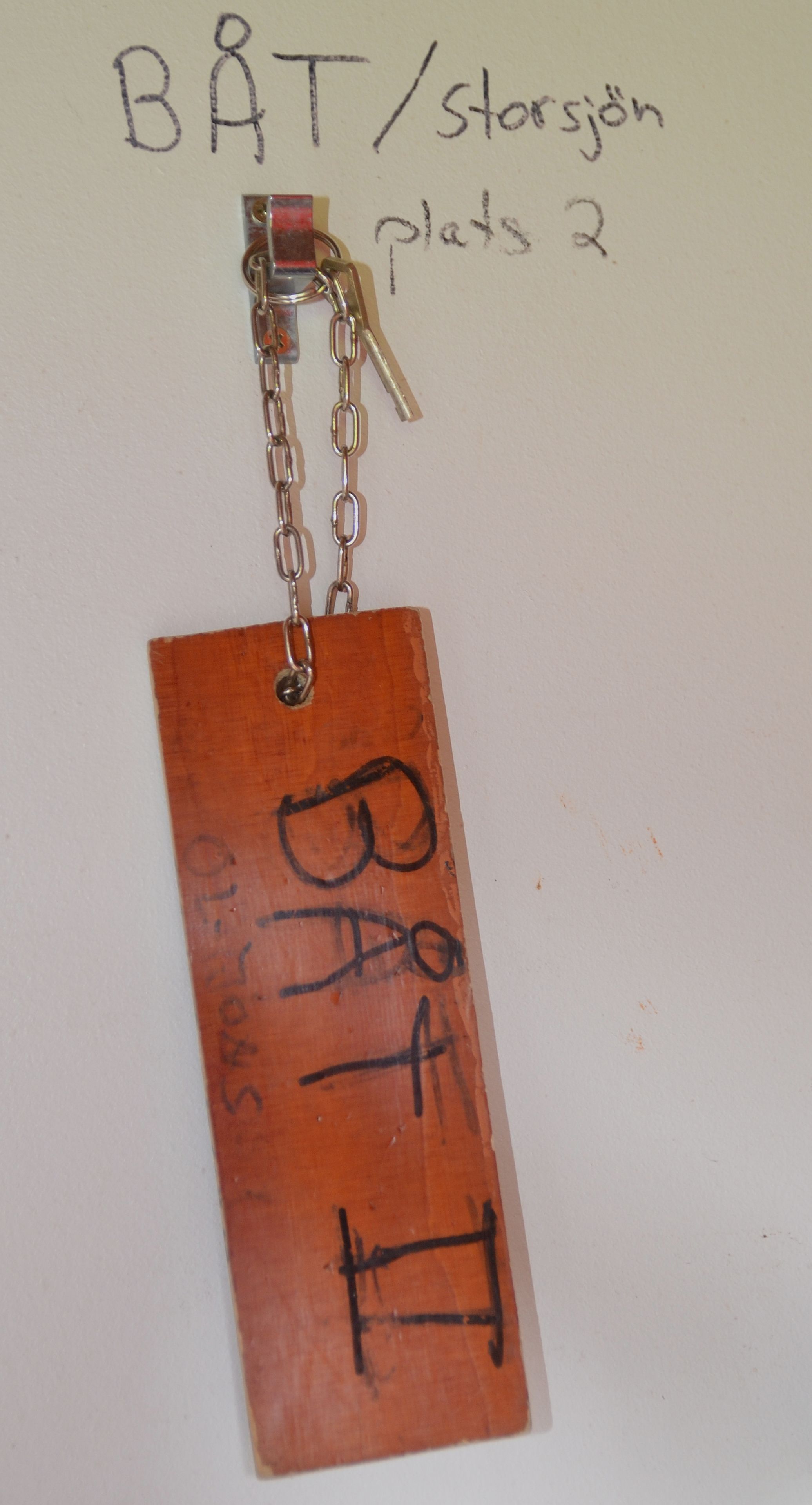 A wooden tag with the text "BAT" and "plats 2" on a white background.