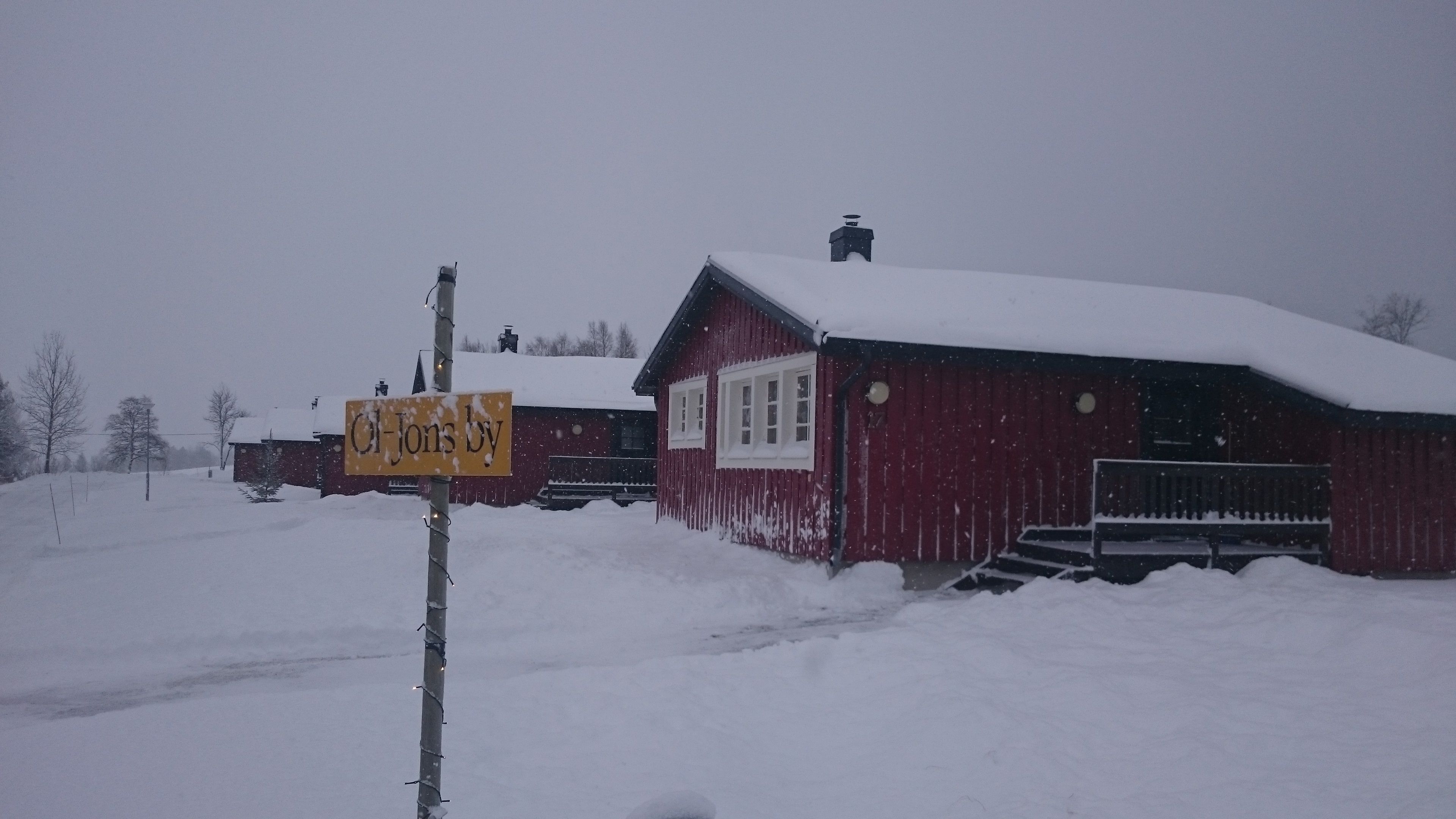 En snötäckt landsortsmiljö med ett rött trähus och en skylt med texten ”O-Jons by”.
