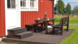 A red building with white trim, a wooden deck with a table and benches, and a small flower vase. - Ol-jons By (Hallen)
