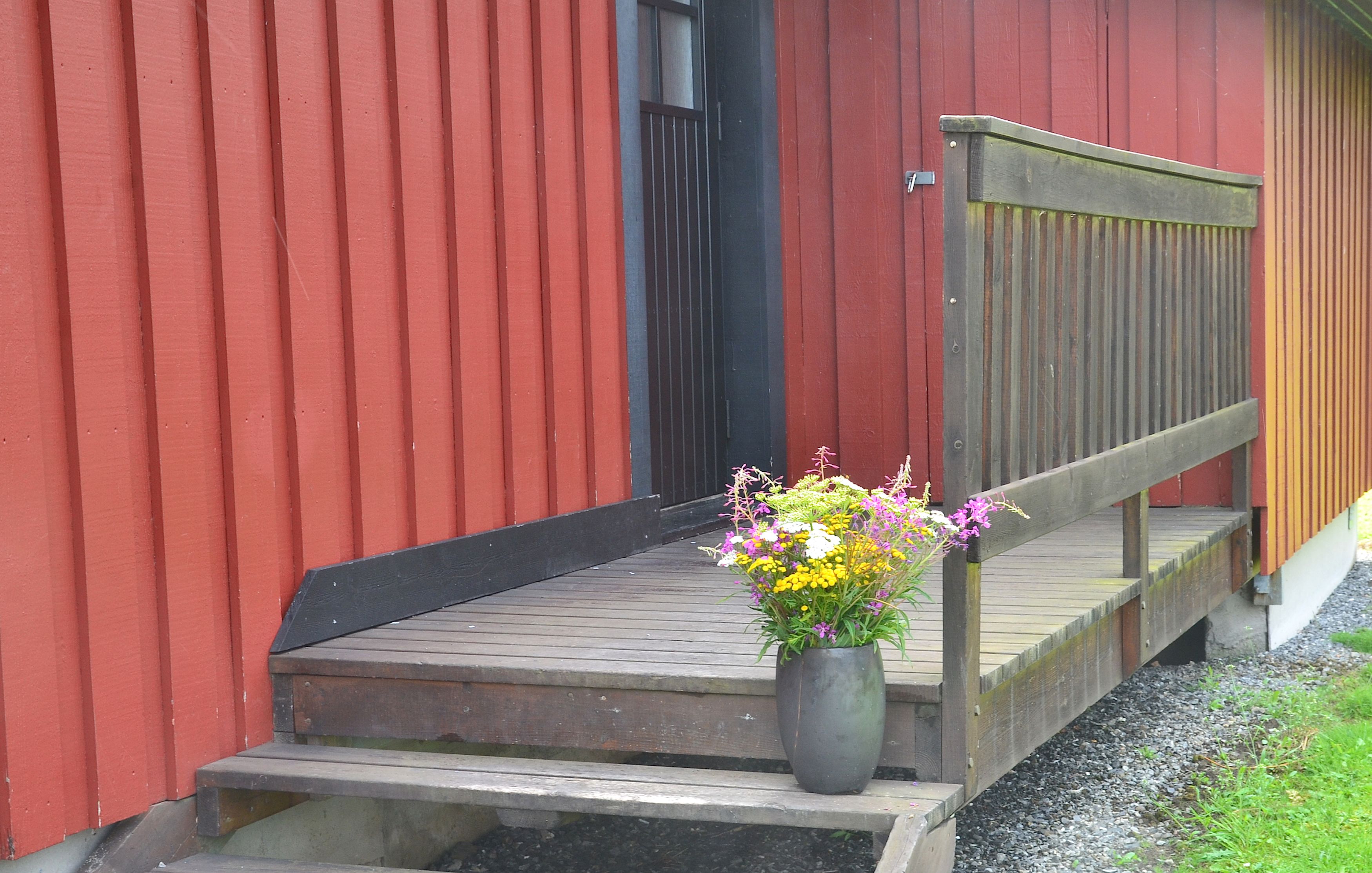 Eine Holzterrasse führt zu einem roten Gebäude mit einer schwarzen Tür. Ein Blumentopf mit Blumen steht auf der Terrasse.