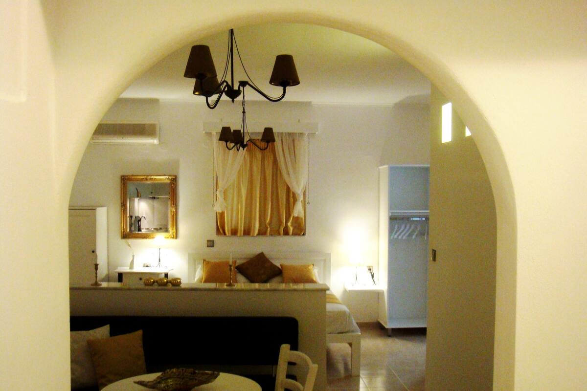 Uma porta arqueada dá acesso a uma sala de jantar com uma mesa redonda e cadeiras, um lustre e uma cama com uma mesinha de cabeceira ao fundo.