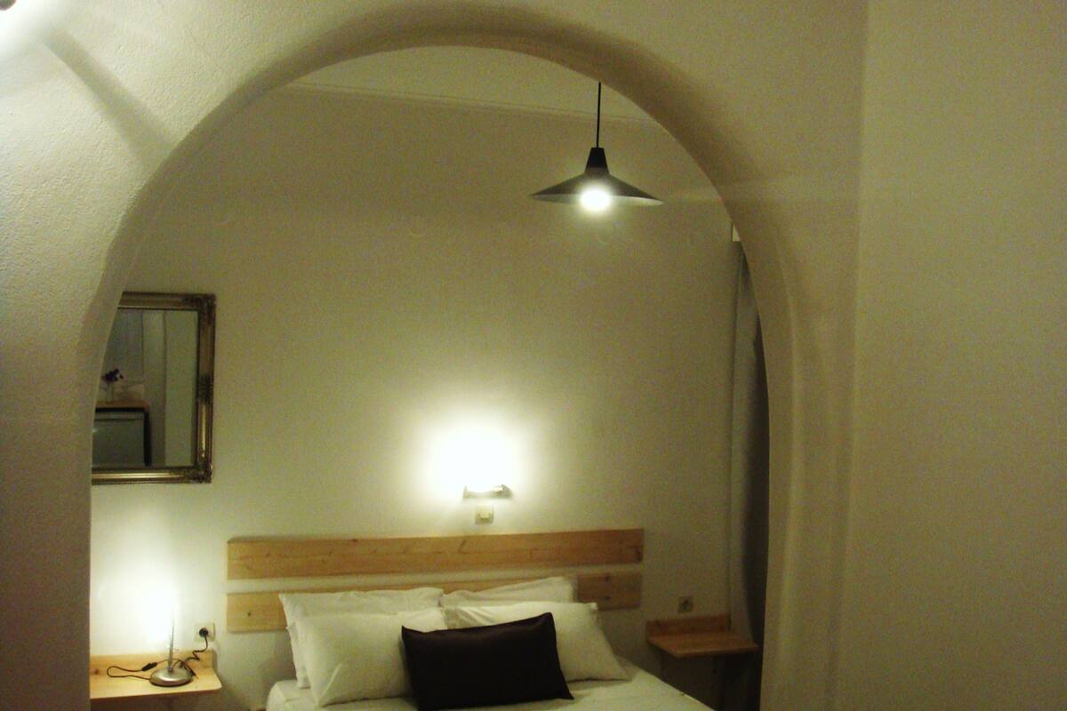 Um quarto com cabeceira de madeira, uma cama de solteiro com almofadas brancas e pretas, uma mesa de cabeceira com abajur e um espelho na parede.