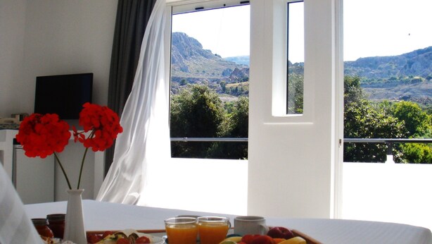 A breakfast tray with fruits, juice, and a plate of eggs on a bed with a view of a mountainous landscape through large windows. - Blugreen Stegna B&B (Rhodes)